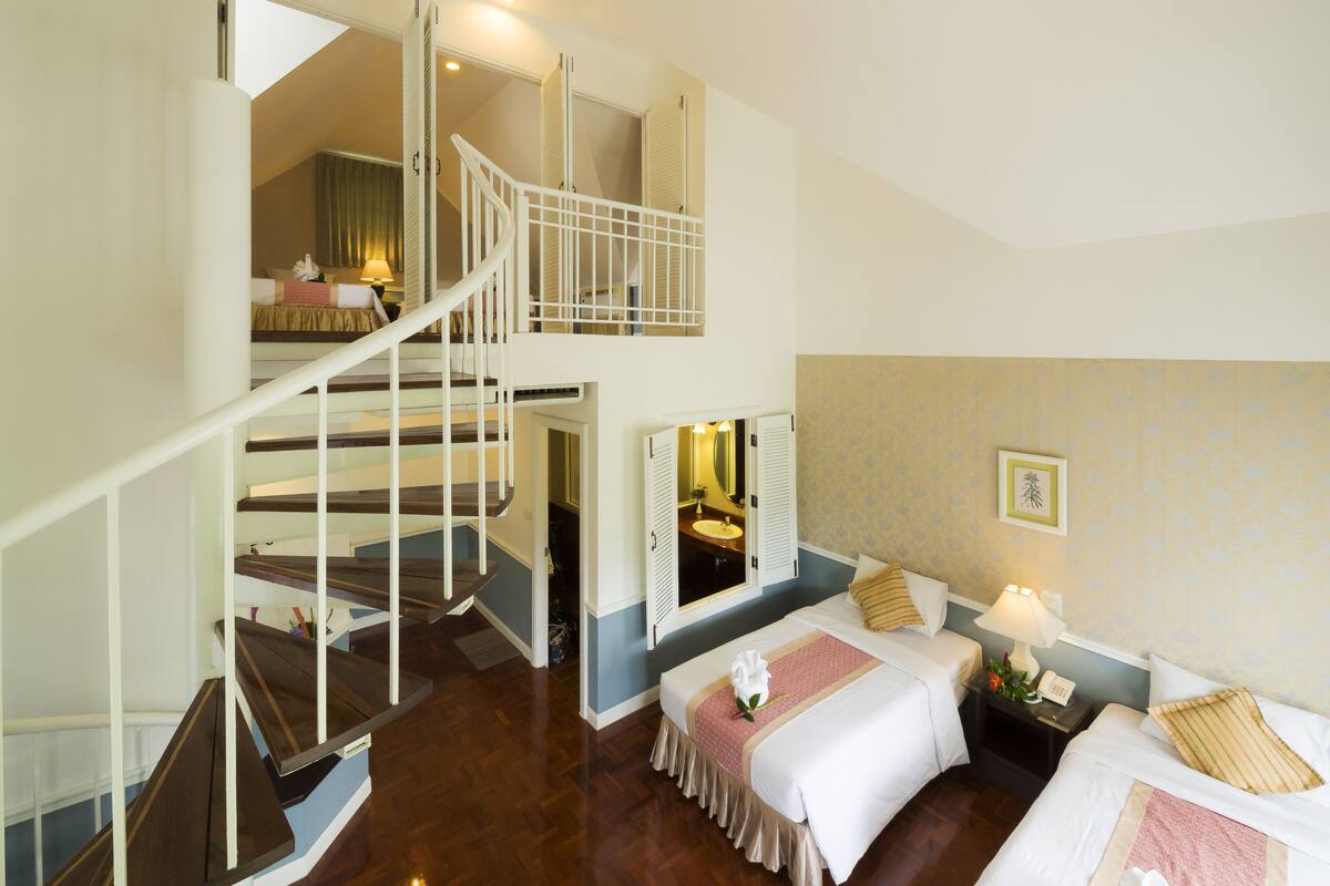 Habitación de hotel con cama doble, un pequeño escritorio con teléfono, mesita de noche con lámpara, baño con espejo y lavamanos, y una escalera que sube a un nivel superior.