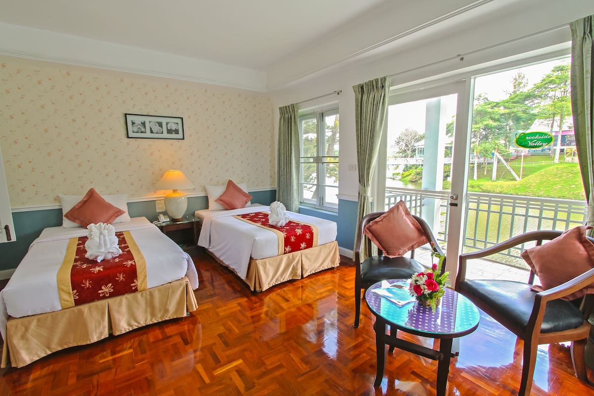 Habitación de hotel con dos camas, una mesita con un jarrón de flores y un balcón con vistas al jardín.