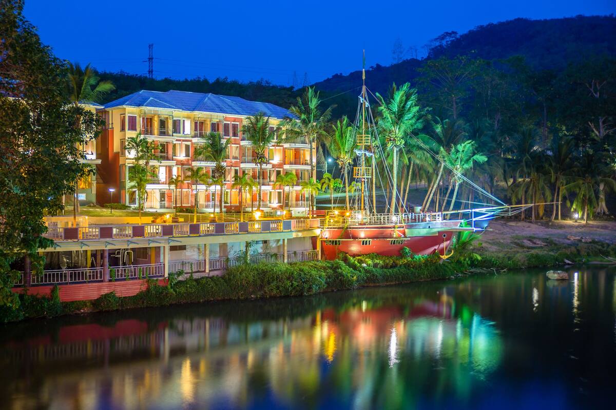 Un hotel frente al agua con varios balcones, un barco amarrado y abundante vegetación.