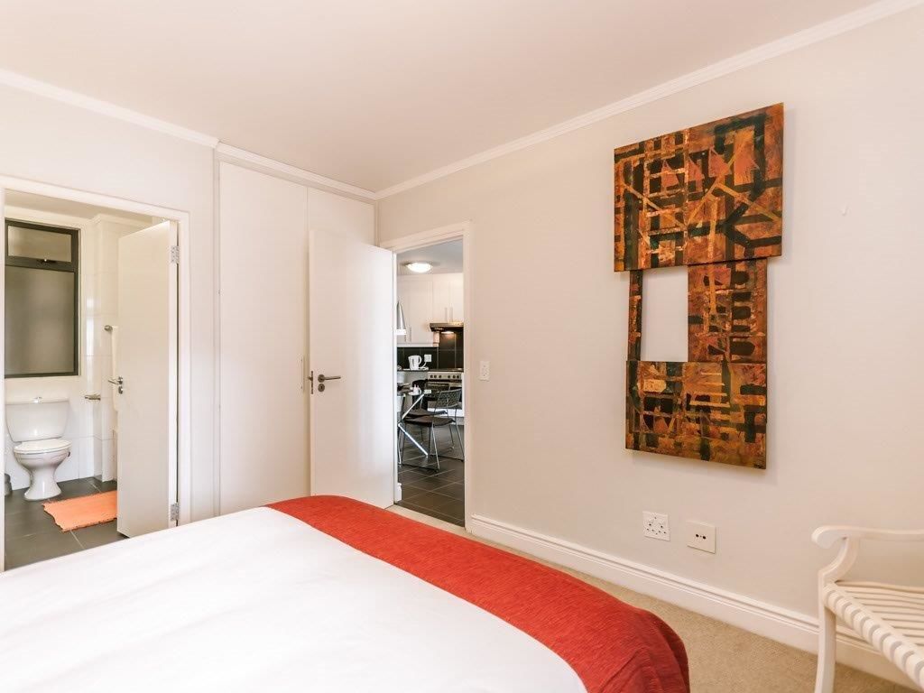 A hotel room with a bed, a red throw, a white chair, and a wall with a geometric art piece.