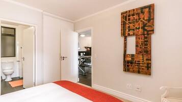 A hotel room with a bed, a red throw, a white chair, and a wall with a geometric art piece.