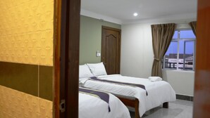 Habitación de hotel con dos camas, una puerta de madera, una ventana con cortinas y vistas a edificios.