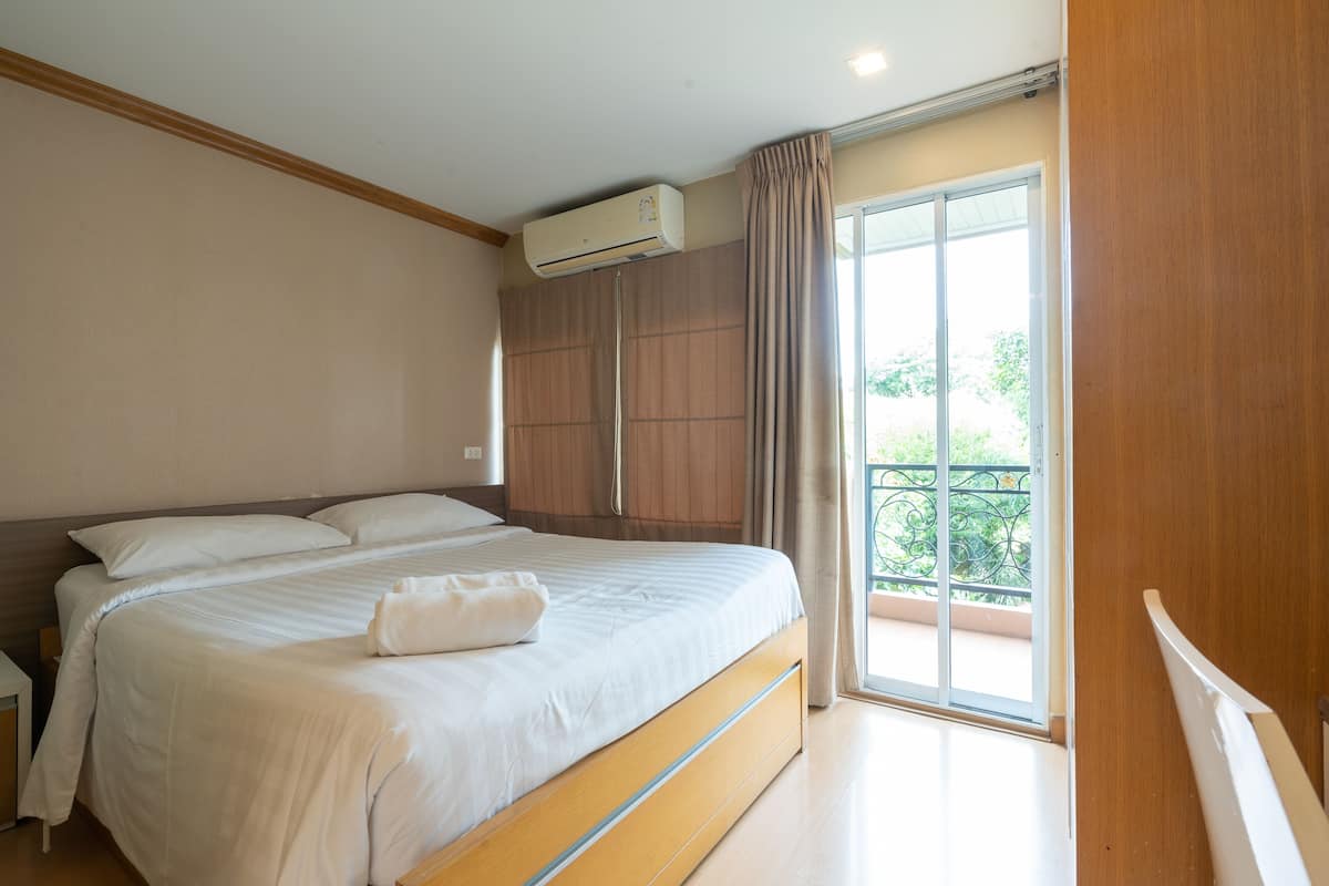 A bedroom with a large bed, an air conditioning unit, a sliding glass door to a balcony, and a wooden floor.