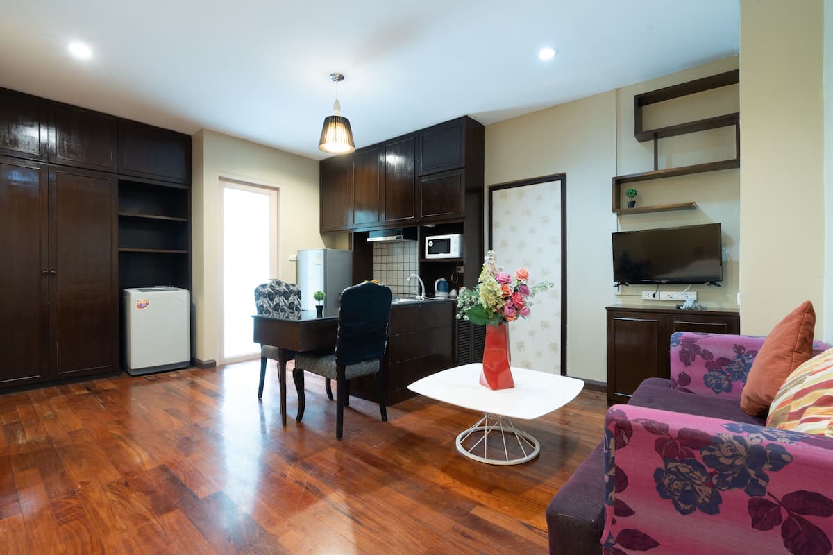 A modern living room with wooden flooring, a dining area, a kitchen with dark cabinets, a white table, and a sofa with floral upholstery.