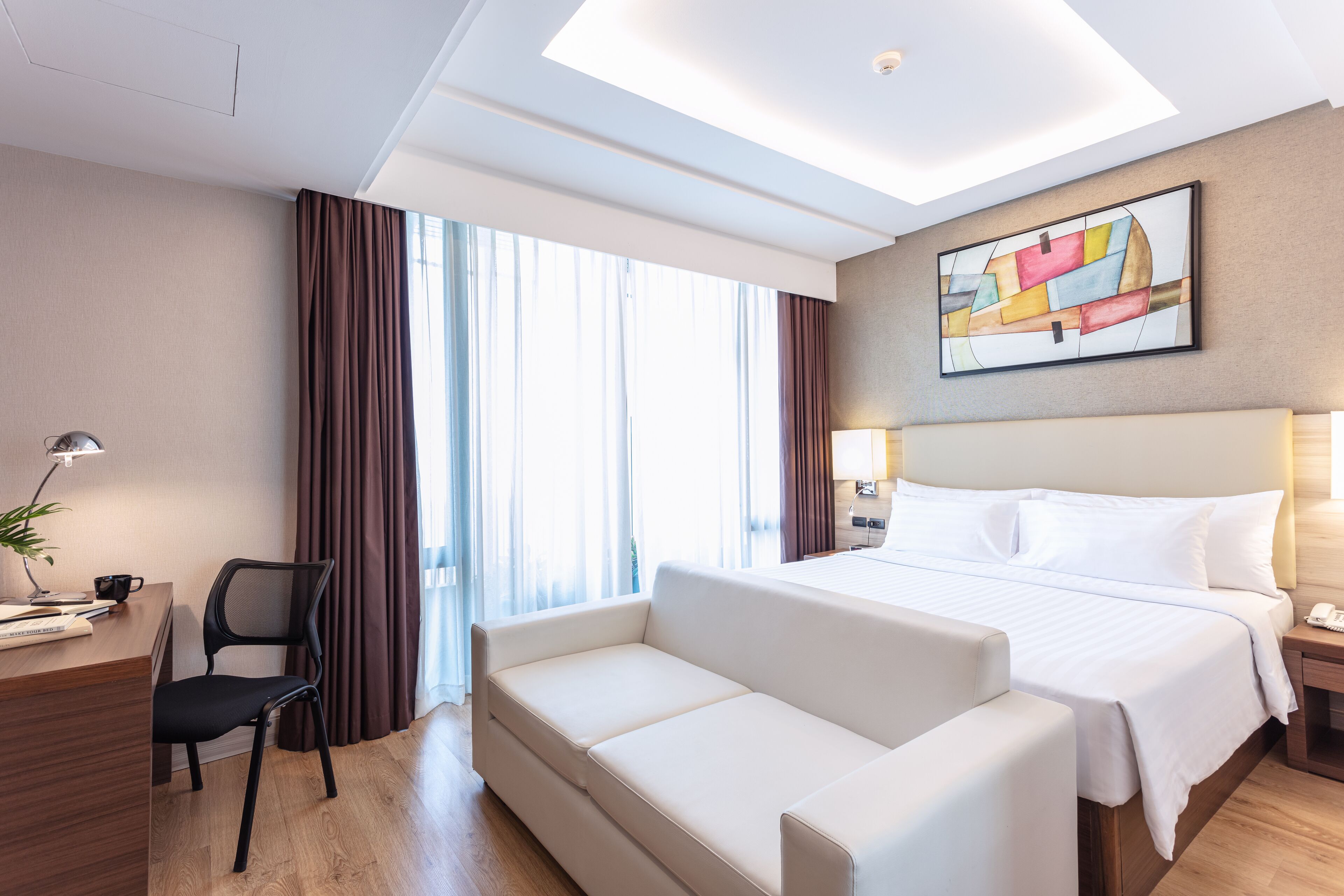 A hotel room with a bed, a desk with a chair, a sofa, a nightstand, and a painting on the wall.