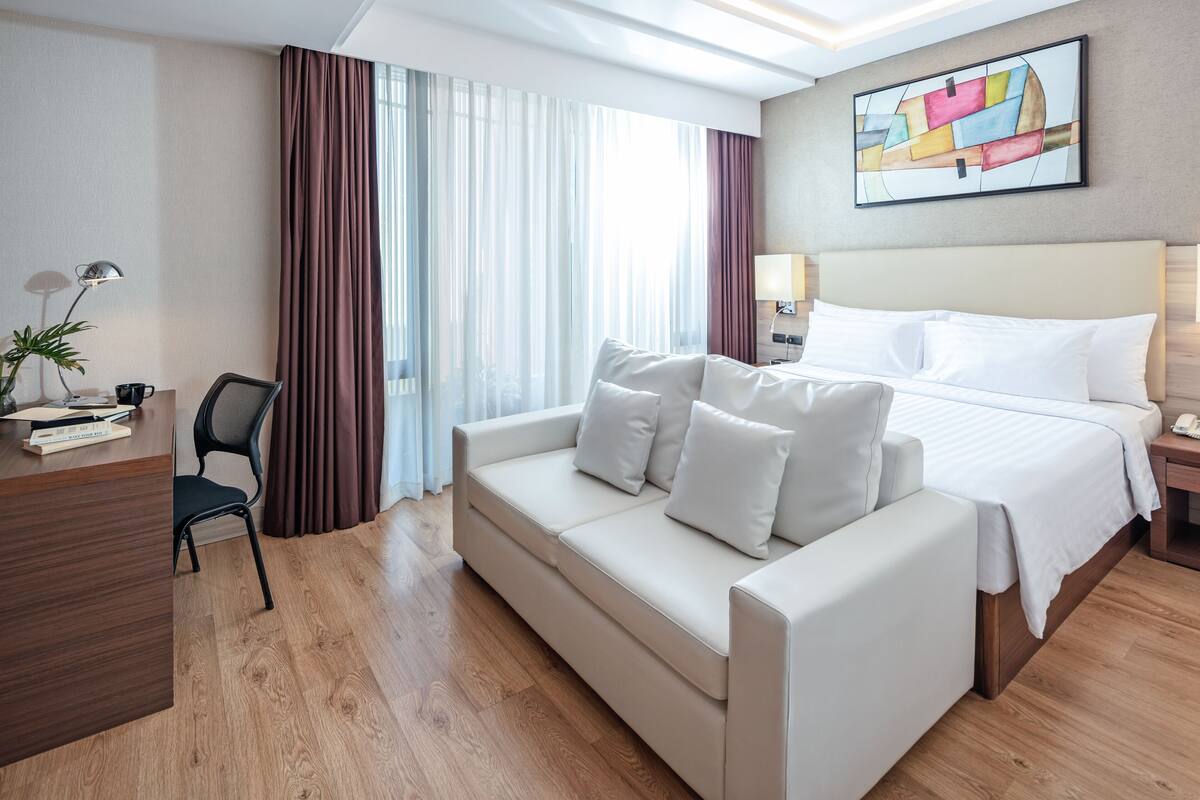 A hotel room with a bed, a sofa, a desk with a lamp, and a window with curtains.
