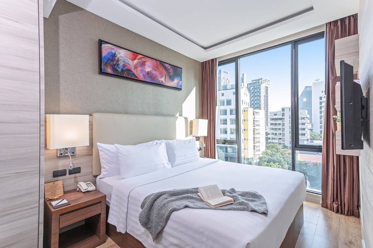 A modern hotel room with a large bed, a flat-screen TV, a bedside table with a lamp, and a window with a city view.