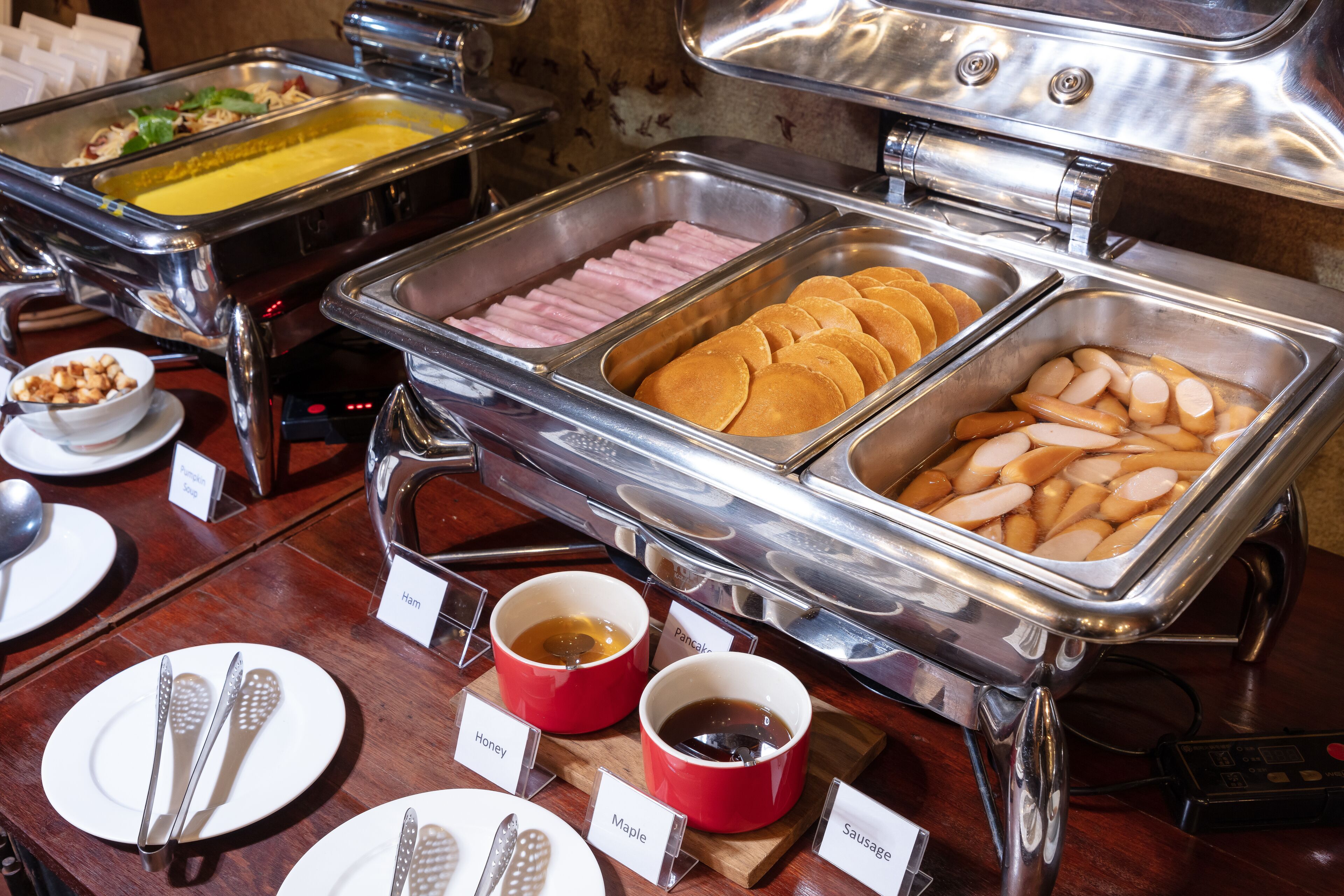 A buffet with labeled containers of various food items such as ham, pancakes, sausages, and maple syrup.