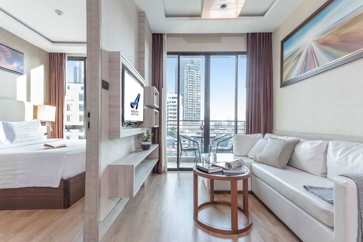 A modern hotel room with a bed, a sofa, a TV, a small table, and a balcony with a city view.