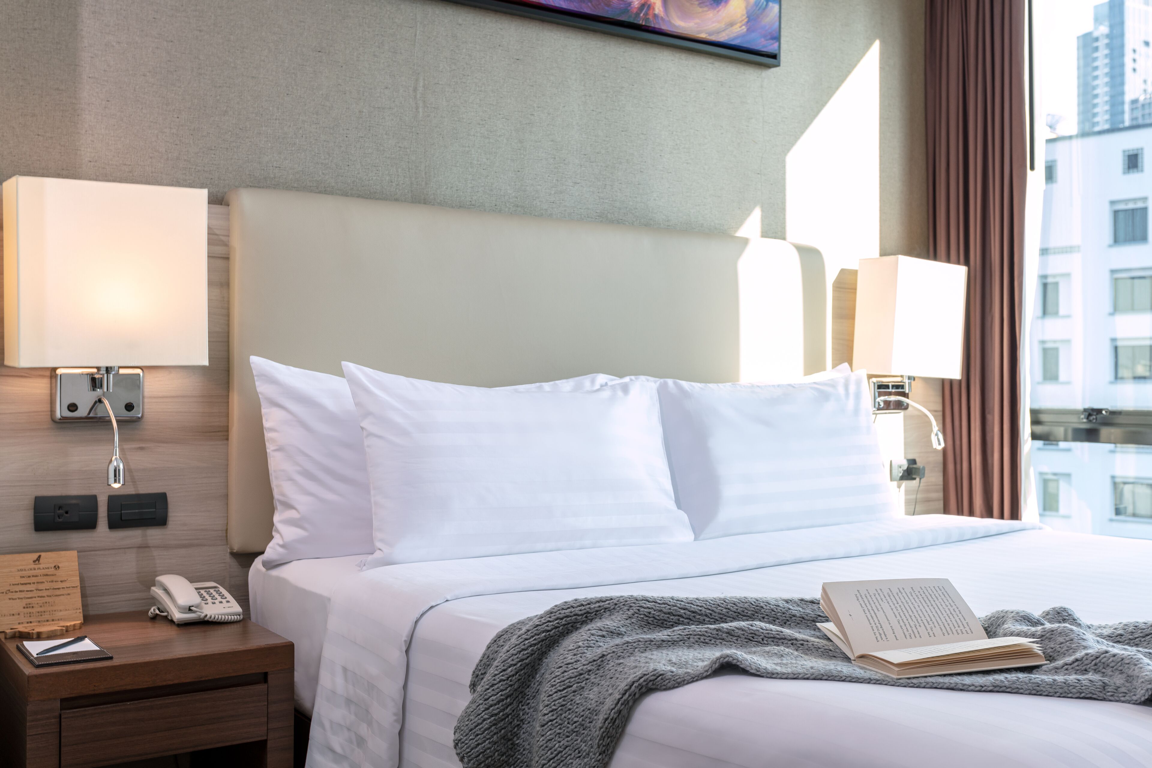 Habitación de hotel con cama, lámparas de noche, teléfono, libreta y un libro sobre la cama.