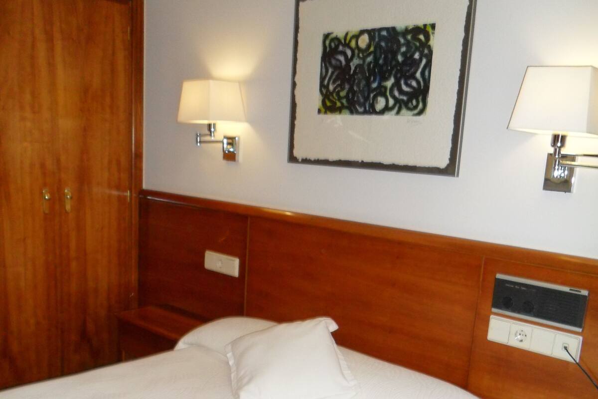 A hotel room with a large bed, wooden headboard, two bedside tables, a nightstand, a wall-mounted lamp, and a framed abstract artwork.