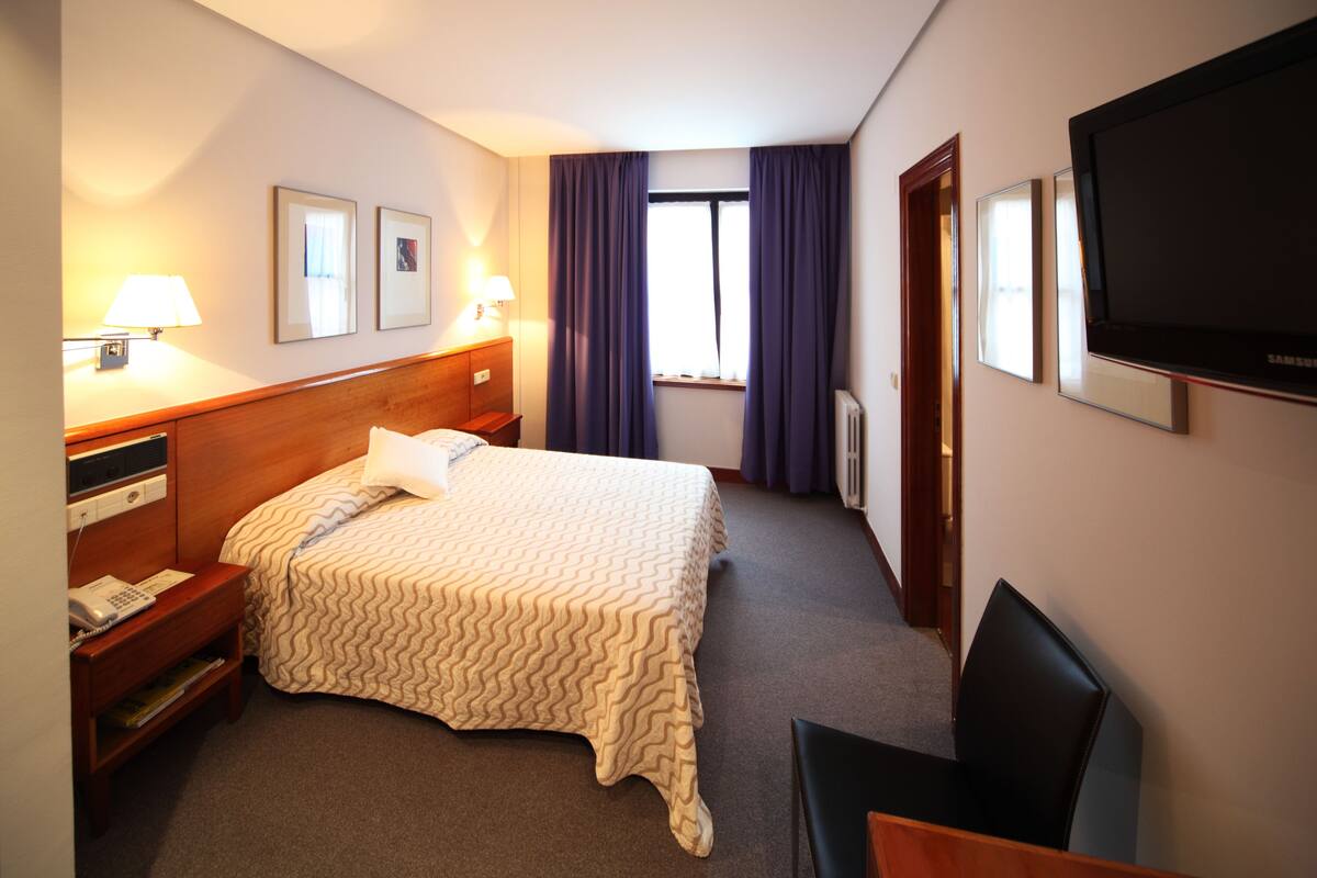 A hotel room with a bed, bedside tables, a desk, a chair, a TV, and a window with curtains.