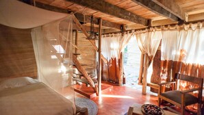 Una habitación rústica de madera con una cama, una silla, una mesita con un frutero y una escalera que conduce a un área exterior.