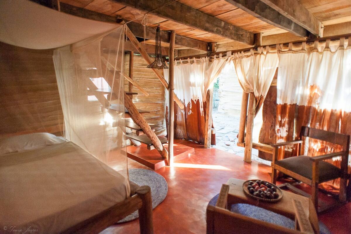 A rustic wooden room with a bed, a chair, a small table with a bowl of fruit, and a staircase leading to an outdoor area.