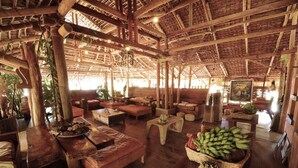 Un salón amplio y rústico con mobiliario de madera y techo de paja, que incluye una gran mesa con fruteros y una lámpara colgante.