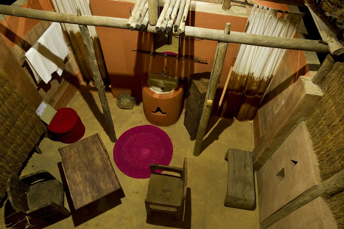 Una habitación rústica con una silla de madera, una mesita pequeña, un tapete tejido rosa, un taburete de madera y un banco de madera.