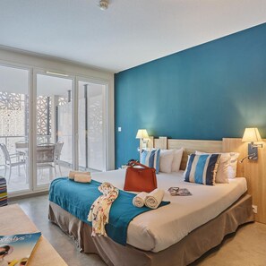A hotel room with a bed, two bedside lamps, a view of the outside, and a table with a magazine. - Hotel Appart'hotel Odalys Nakara (Cap d'Agde)