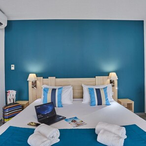 A hotel room with a bed, two bedside lamps, a laptop, a smartphone, a set of books, and a blue wall.