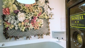 Pared decorada con diversas flores, un letrero que dice "HOSTAL VAZQUEZ DE MELLA" y un baño con lavamanos y espejo.