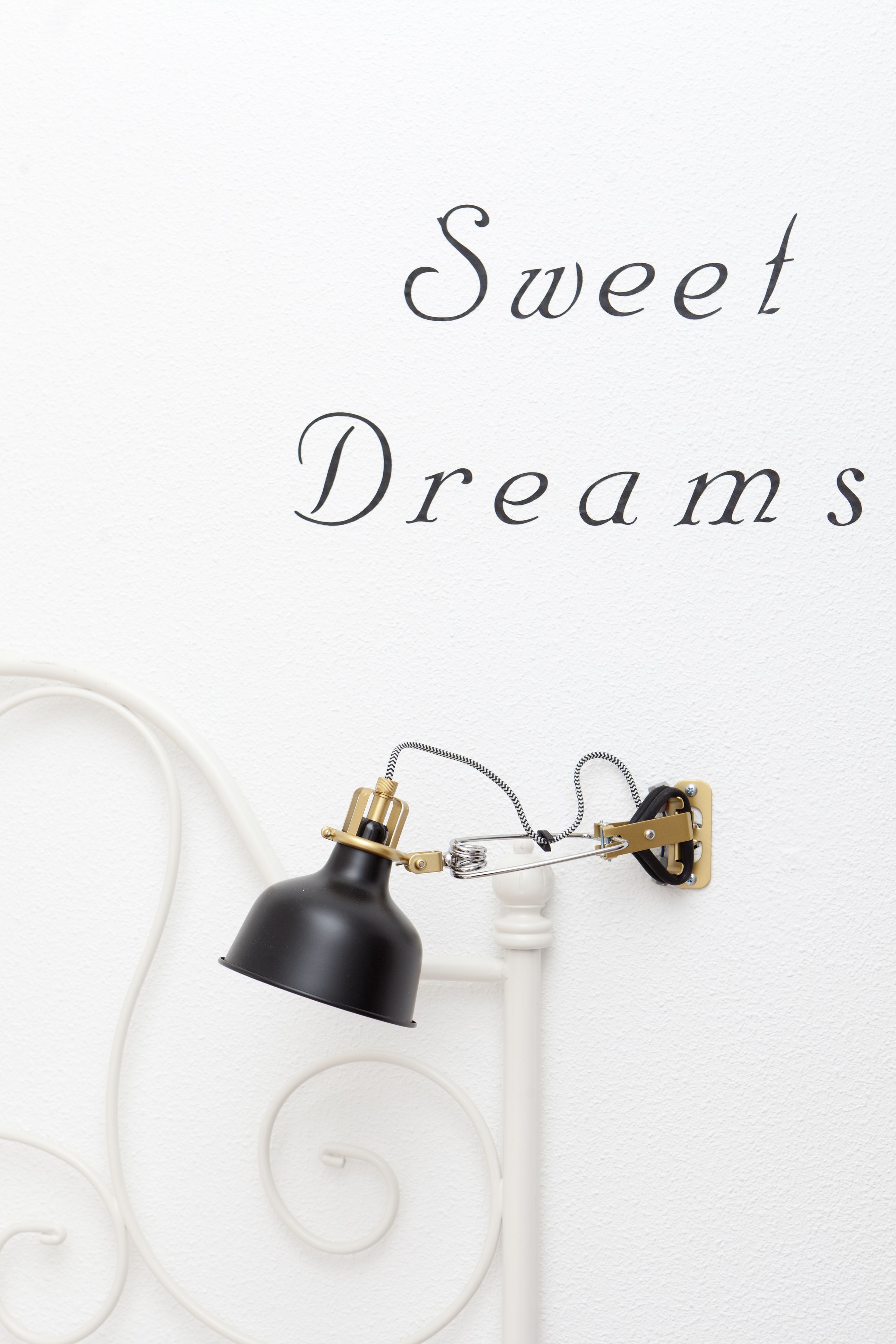 A wall-mounted lamp with a black shade and brass accents, positioned next to a white wall with the text "Sweet Dreams" in a cursive font.