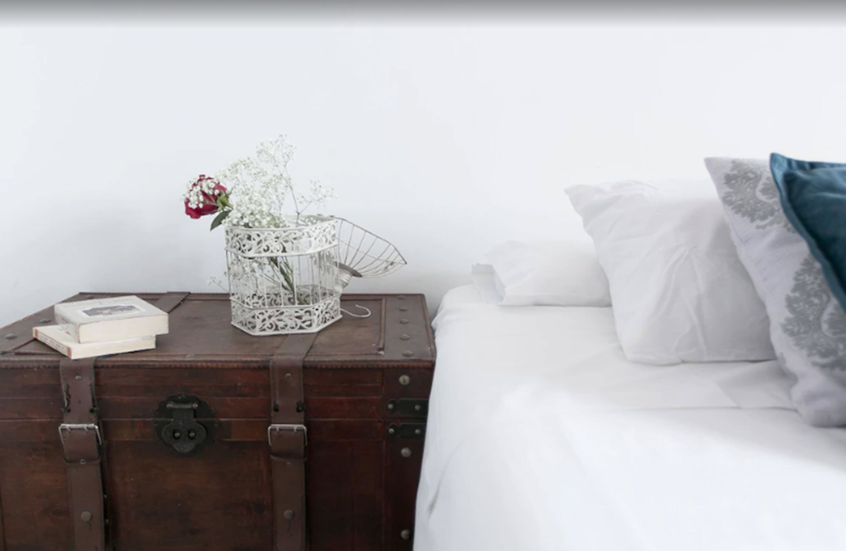 A bed with white bedding and a pillow with a gray pattern, a white vase with flowers, and a book on a wooden chest.