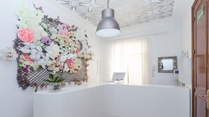 Un área de recepción con un mostrador blanco, un arreglo floral decorativo, una lámpara en el techo y una puerta con la leyenda 'SALIDA'.
