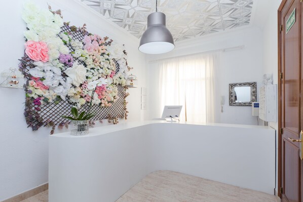 Un área de recepción con un mostrador blanco, un arreglo floral decorativo, una lámpara en el techo y una puerta con la leyenda 'SALIDA'.