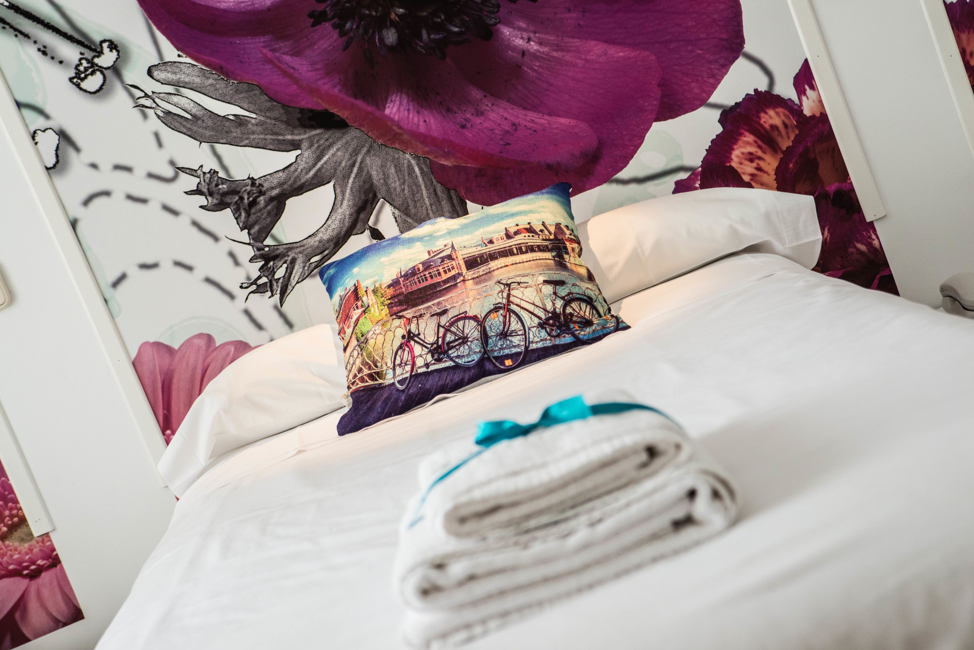 A neatly made bed with a pillow featuring a cityscape and a bicycle, a folded towel, and a decorative headboard with a large purple flower.