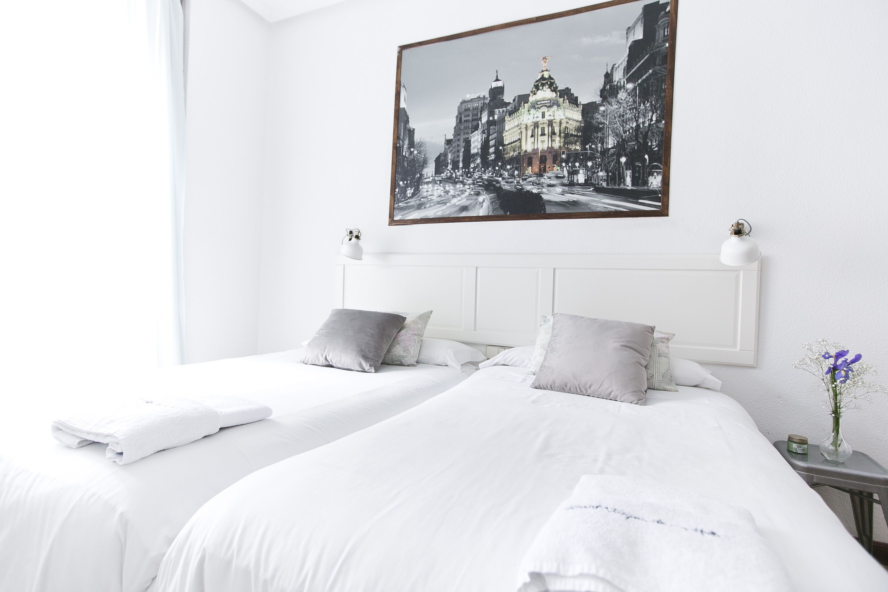 A hotel room with two beds, white bedding, a framed cityscape picture on the wall, and a small table with a vase of flowers.