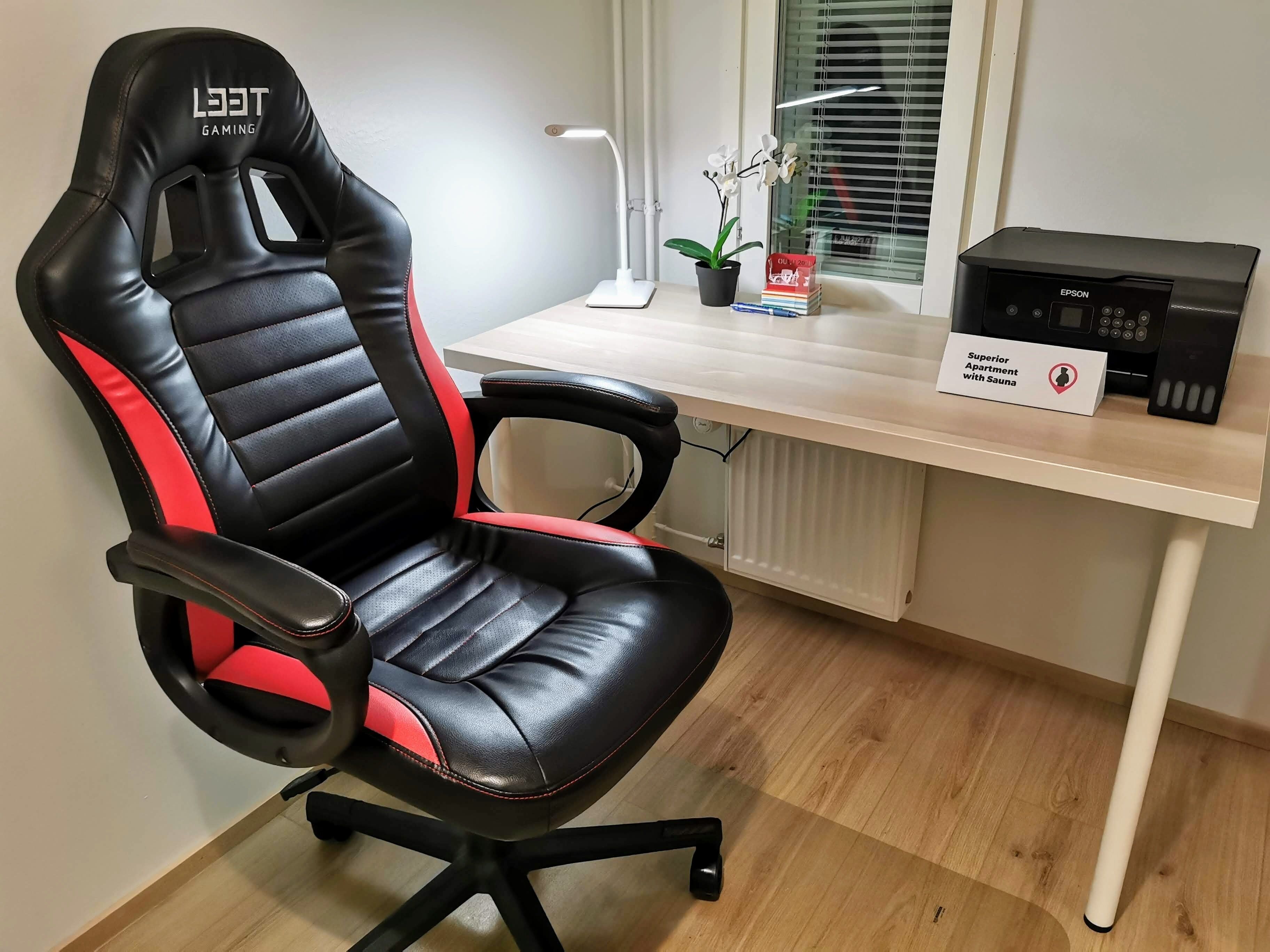 A gaming chair with a red and black design, a desk with a lamp, a potted plant, and an Epson printer.