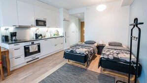 Habitación compacta y moderna con kitchenette, cama con cubrecama estampado, mesita de noche con lámpara y estante montado en la pared.