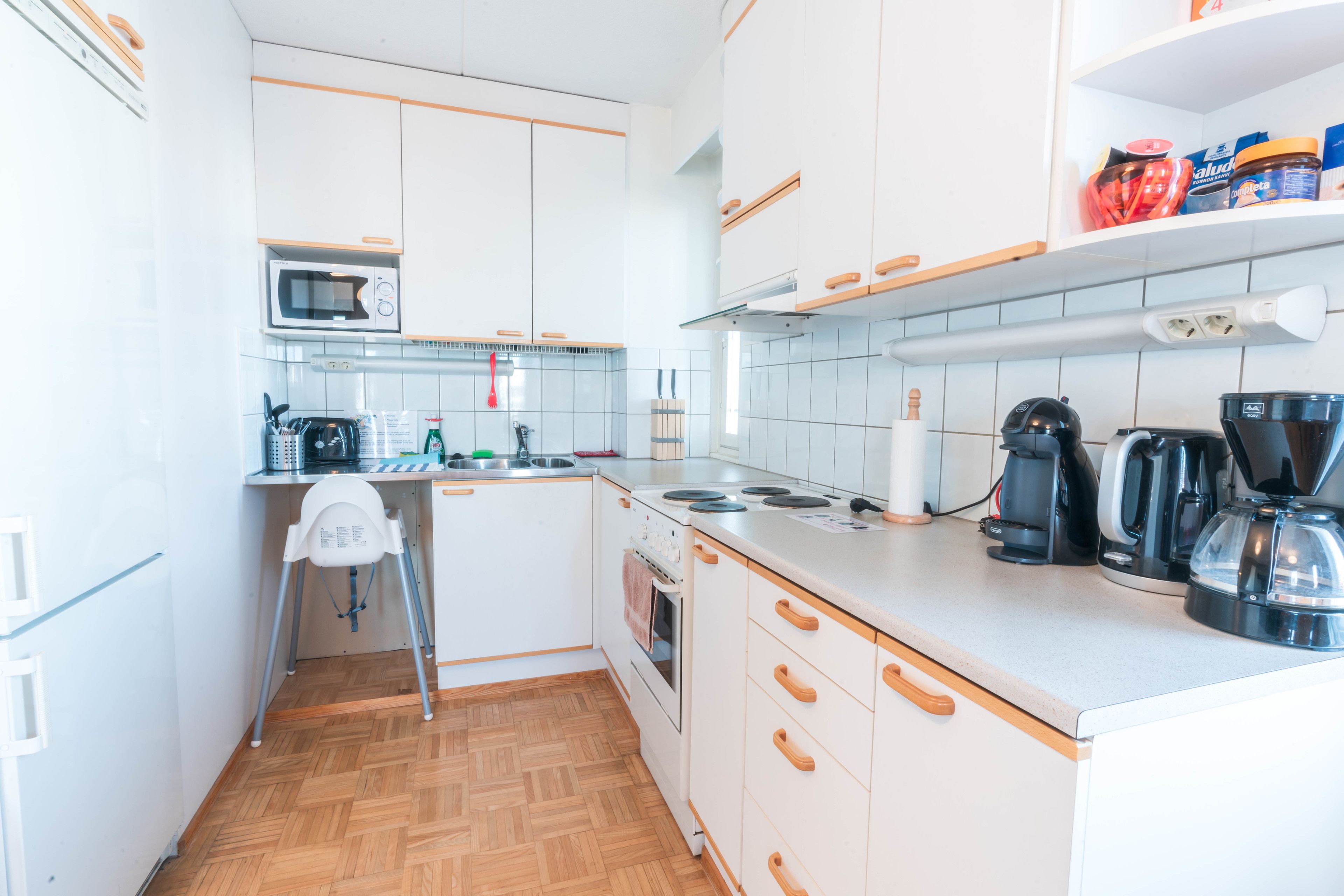 Een kleine keuken met witte kastjes, een magnetron, waterkoker en koffiezetapparaat.