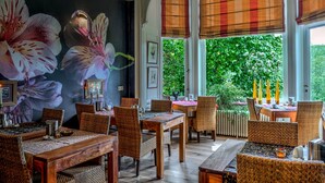 A dining area with wicker furniture, a large floral mural, and a view of greenery outside. - Hotel Dufays (Stavelot)