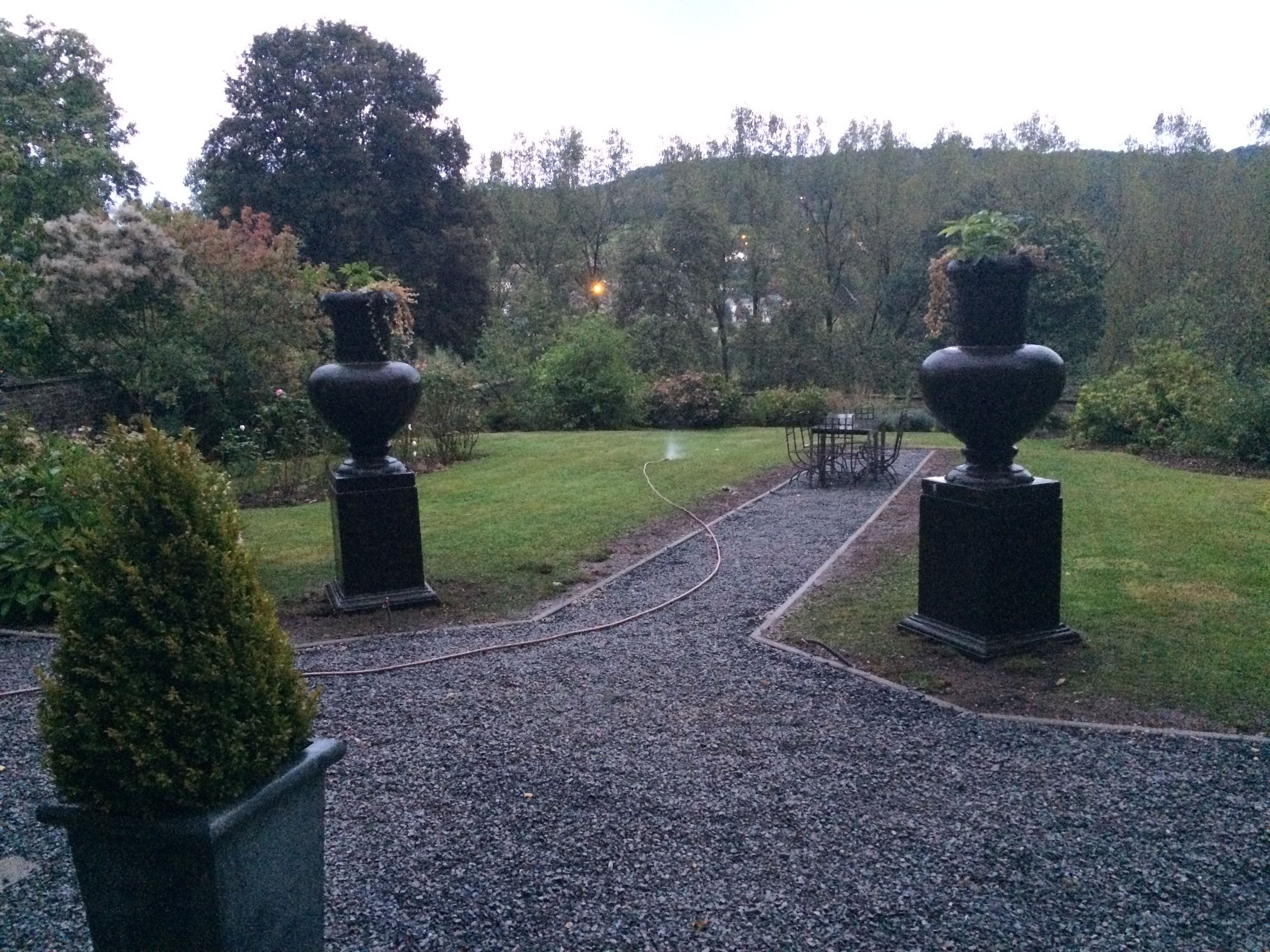 Un chemin de jardin bordé de deux grands vases posés sur des piédestaux, le tout entouré de verdure et d’une pelouse.