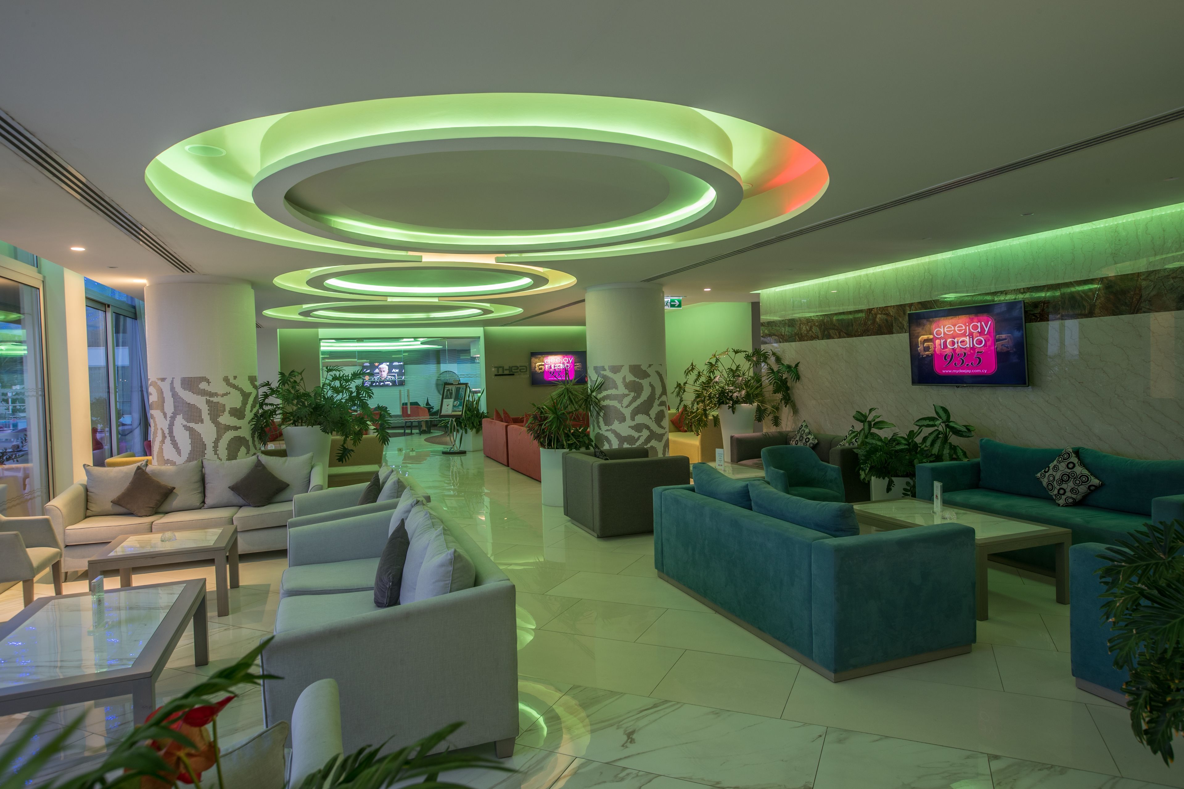 Een moderne lobby met ronde plafondlampen, comfortabele zitplaatsen en een televisiescherm waarop een radiozender wordt vertoond.