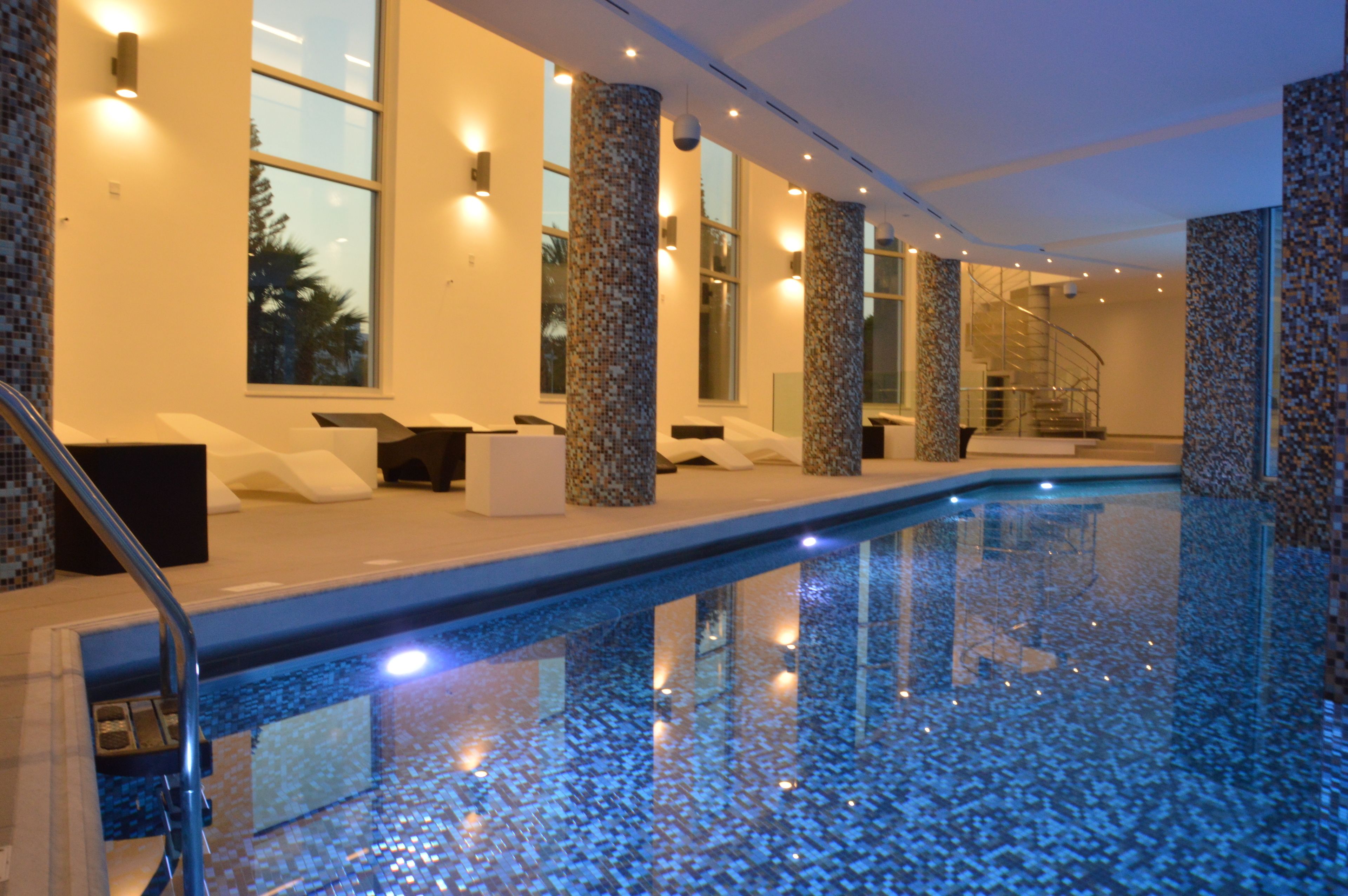 A modern indoor swimming pool with mosaic-tiled walls and a glass railing.