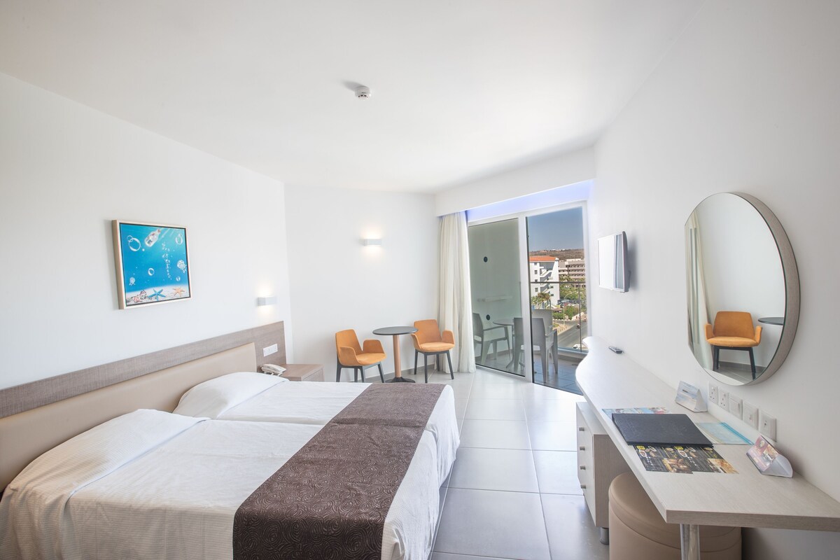Une chambre d’hôtel comprenant un lit, un bureau, une chaise, une petite table, un miroir et une vue sur l’extérieur.