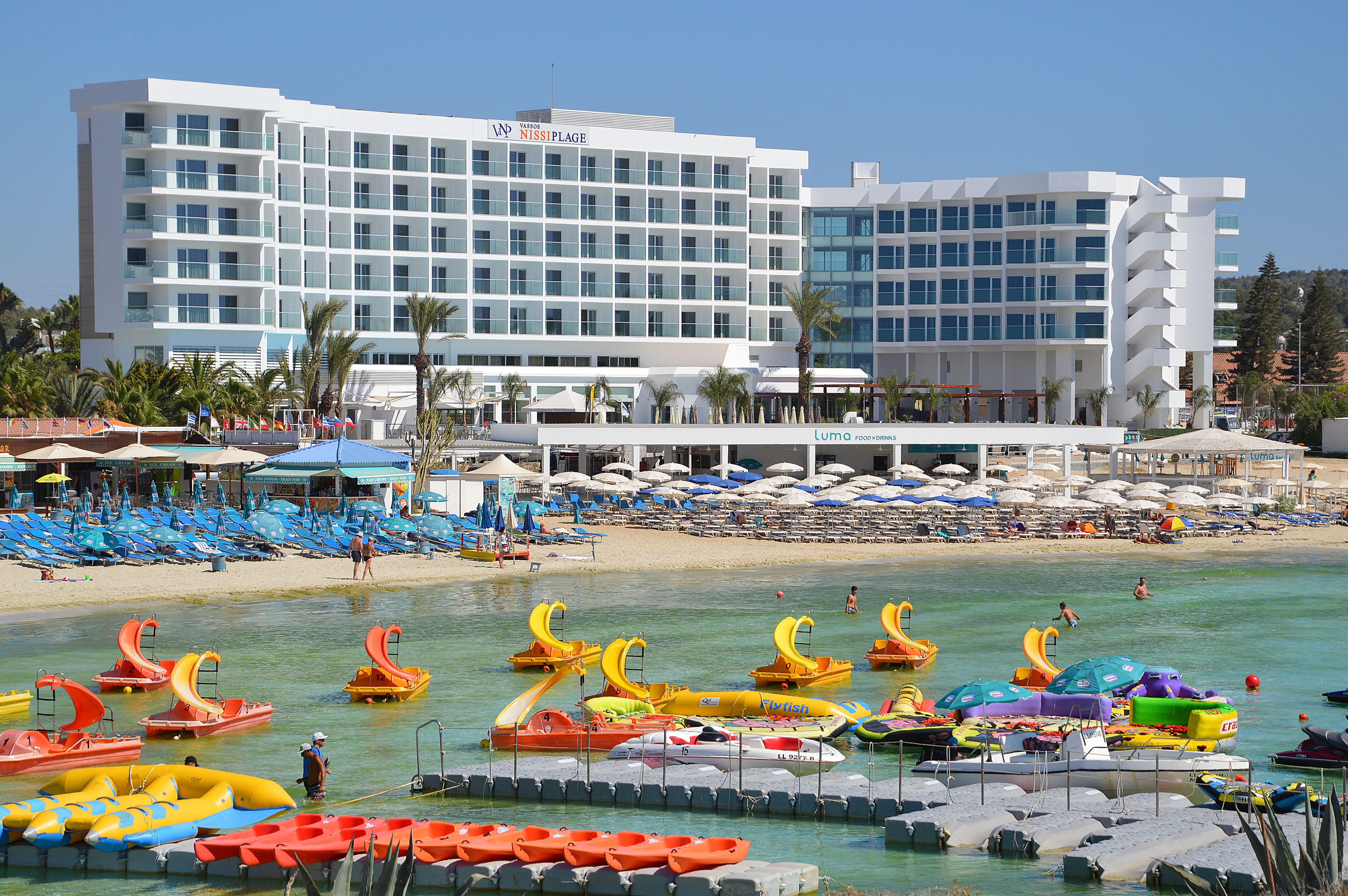 Un hotel con múltiples balcones, una zona de playa con sombrillas y diversos vehículos acuáticos.