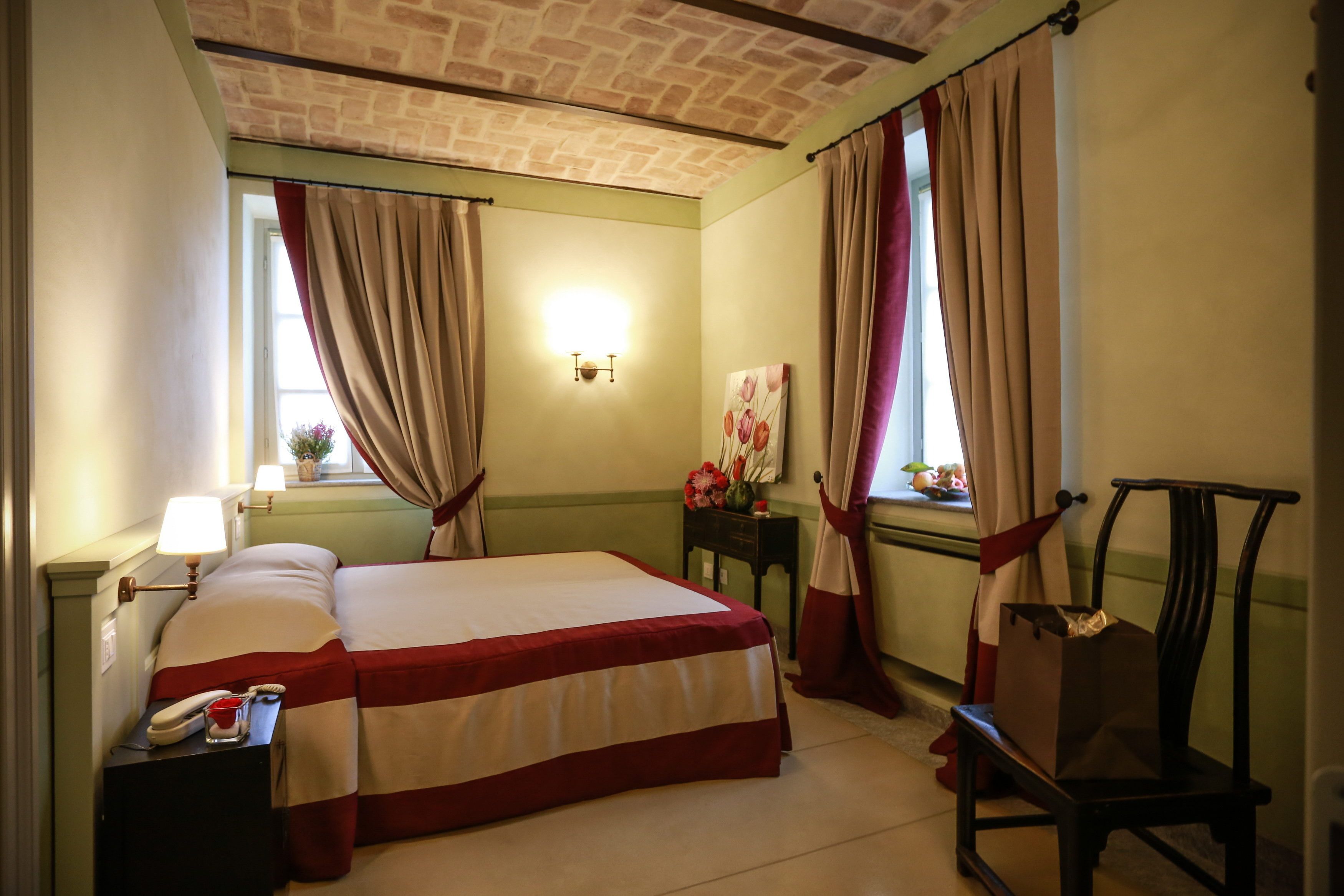 A hotel room with a bed, bedside table with a lamp, a chair, a nightstand with a phone, and a window with curtains.
