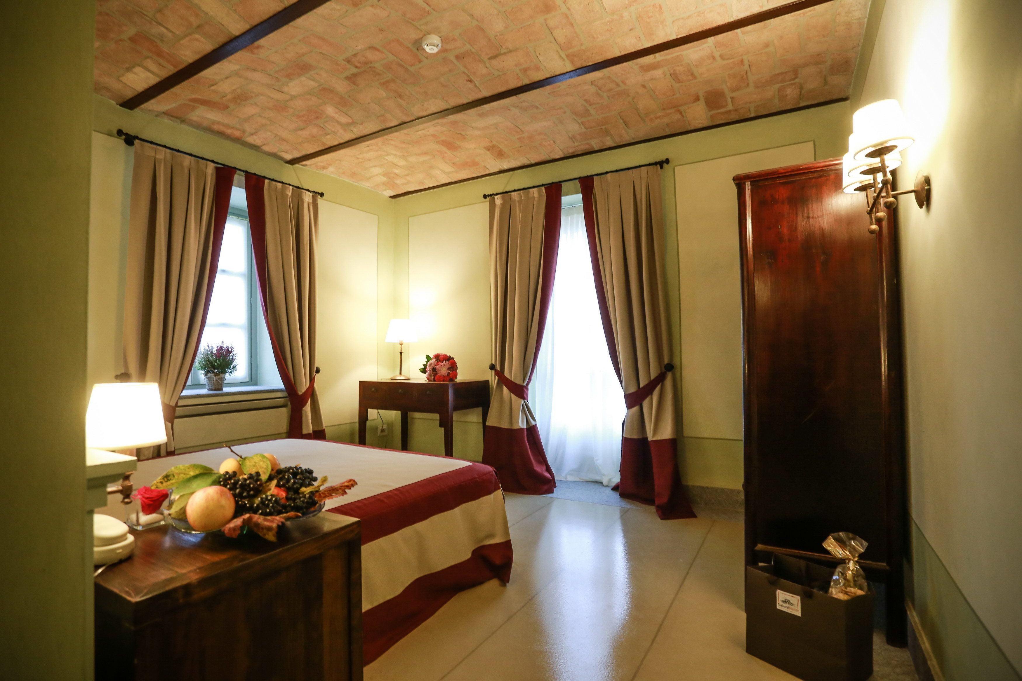 A hotel room with a bed, a desk with a lamp, a small table with a vase, and a trash bin.