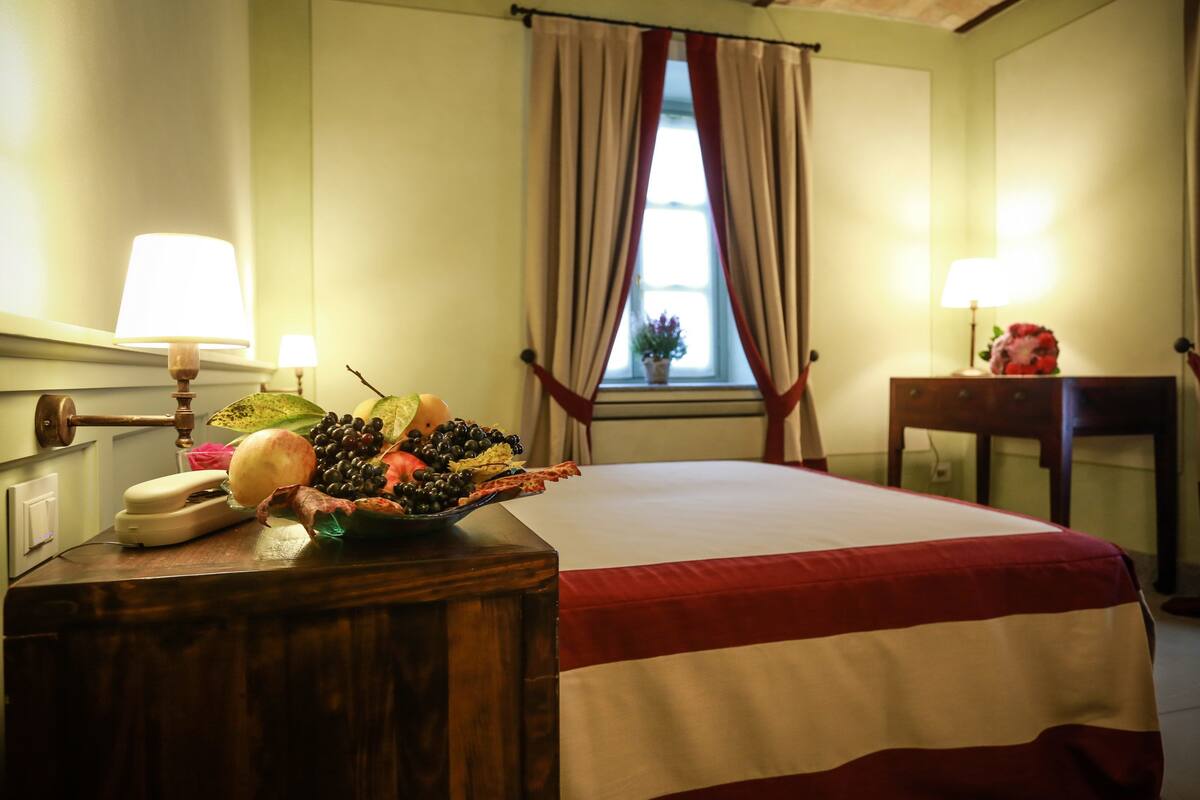 Une chambre d’hôtel avec un lit, une table de chevet en bois avec une lampe, une petite table avec un vase de fleurs et une fenêtre avec des rideaux.