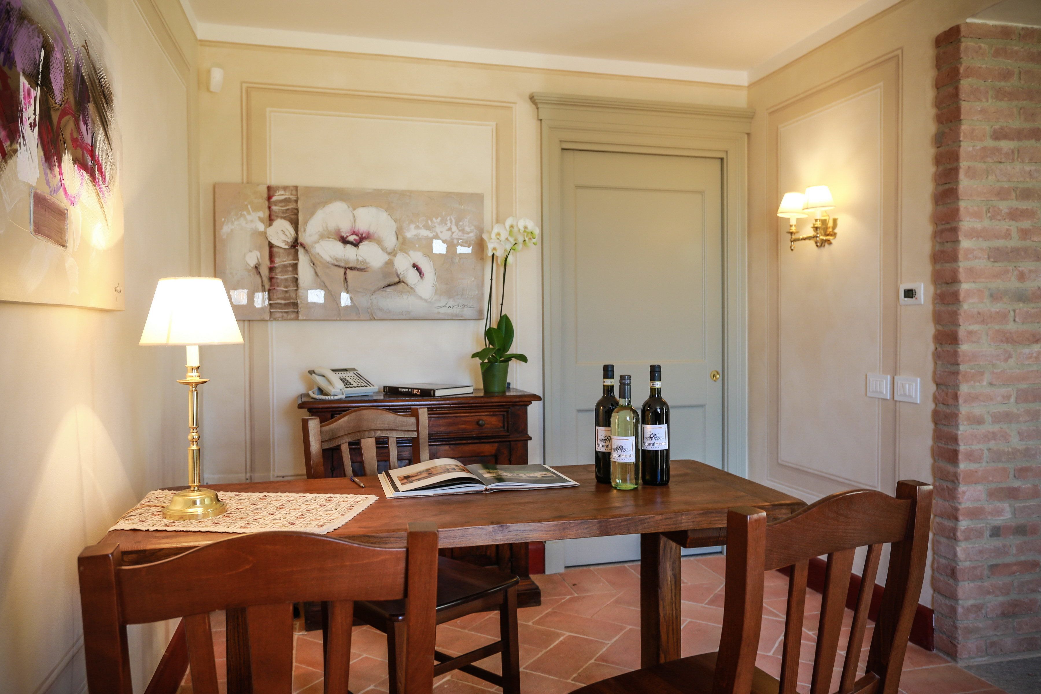 A dining area with a wooden table, chairs, a lamp, a book, and two bottles of wine.