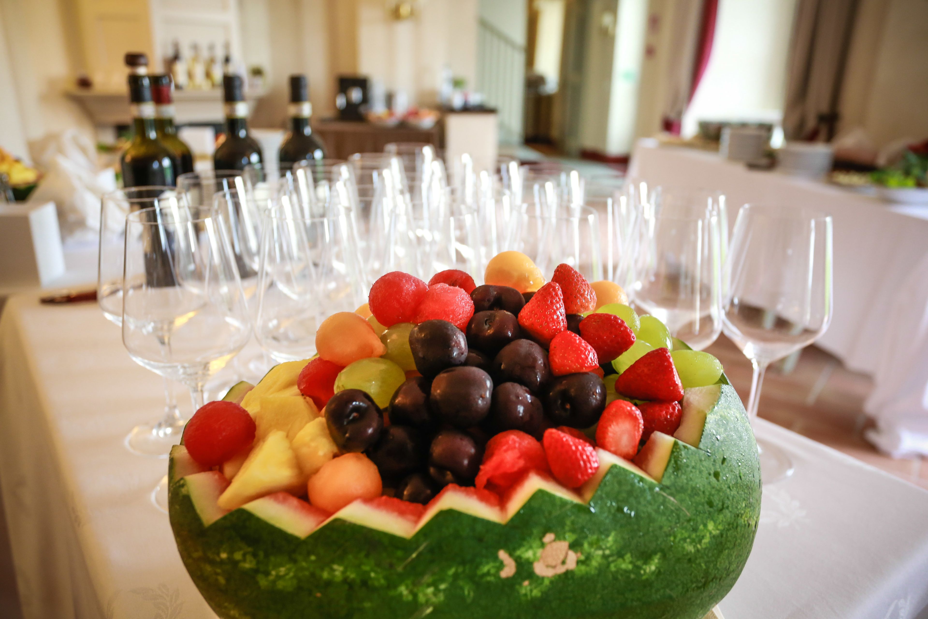 A fruit arrangement on a table with wine glasses and bottles in the background.
