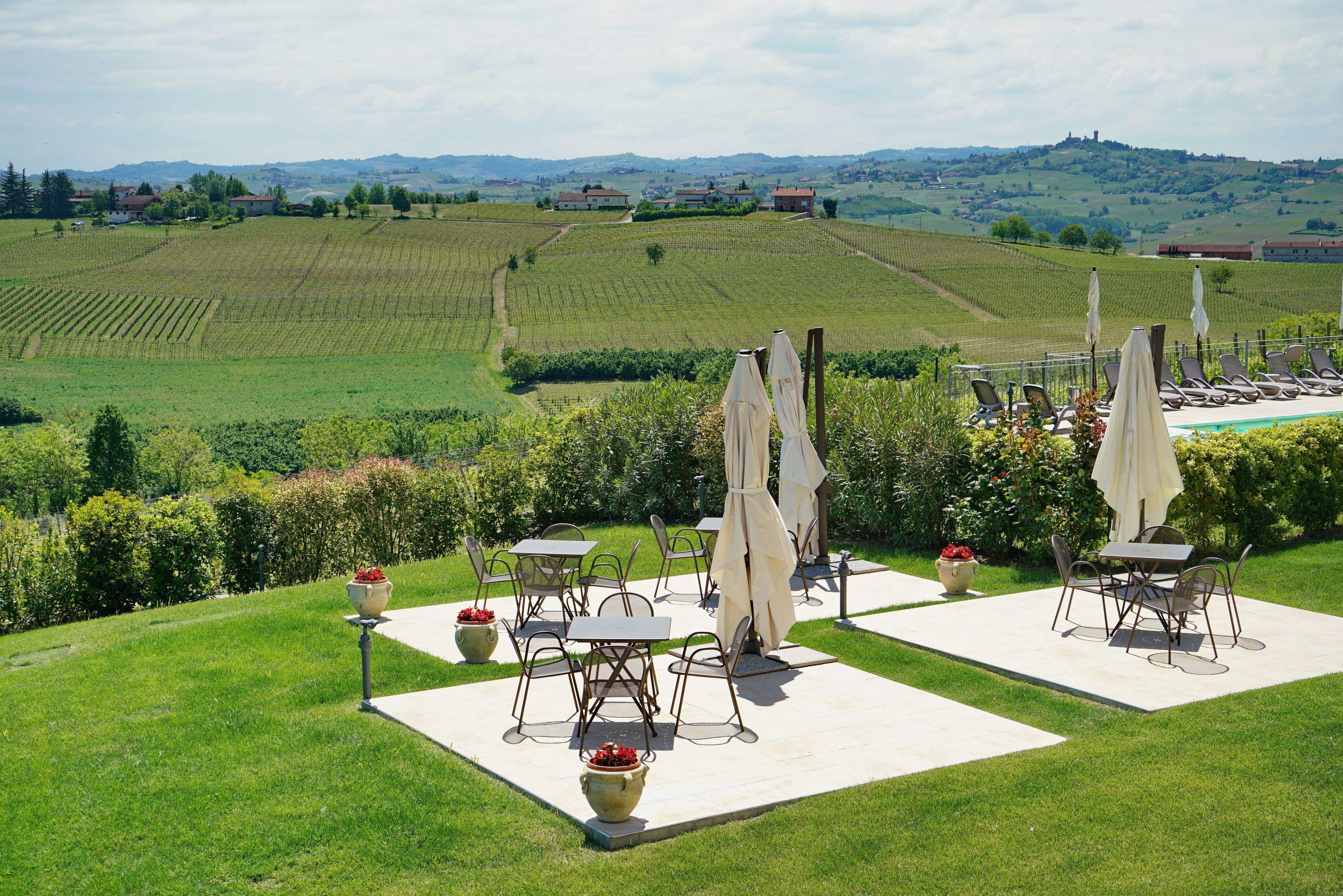 An outdoor patio with tables and chairs, surrounded by a well-maintained lawn and a view of vineyards and hills.