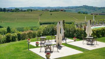 An outdoor patio with tables and chairs, surrounded by a well-maintained lawn and a view of vineyards and hills.