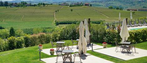 Un patio al aire libre con mesas y sillas, rodeado de un césped cuidado y vistas a viñedos y colinas.