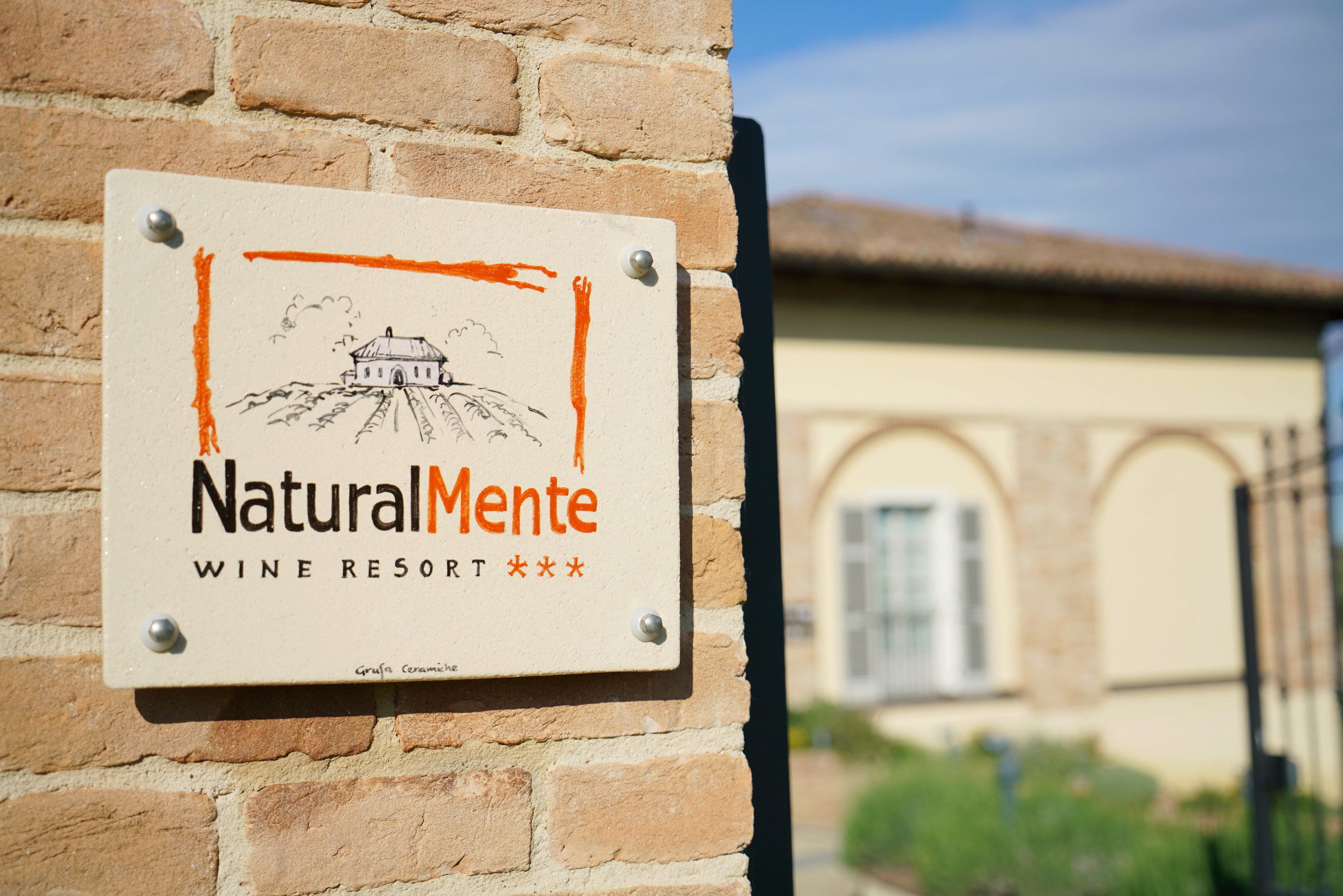 A sign for NaturalMente Wine Resort with a drawing of a building and hills.