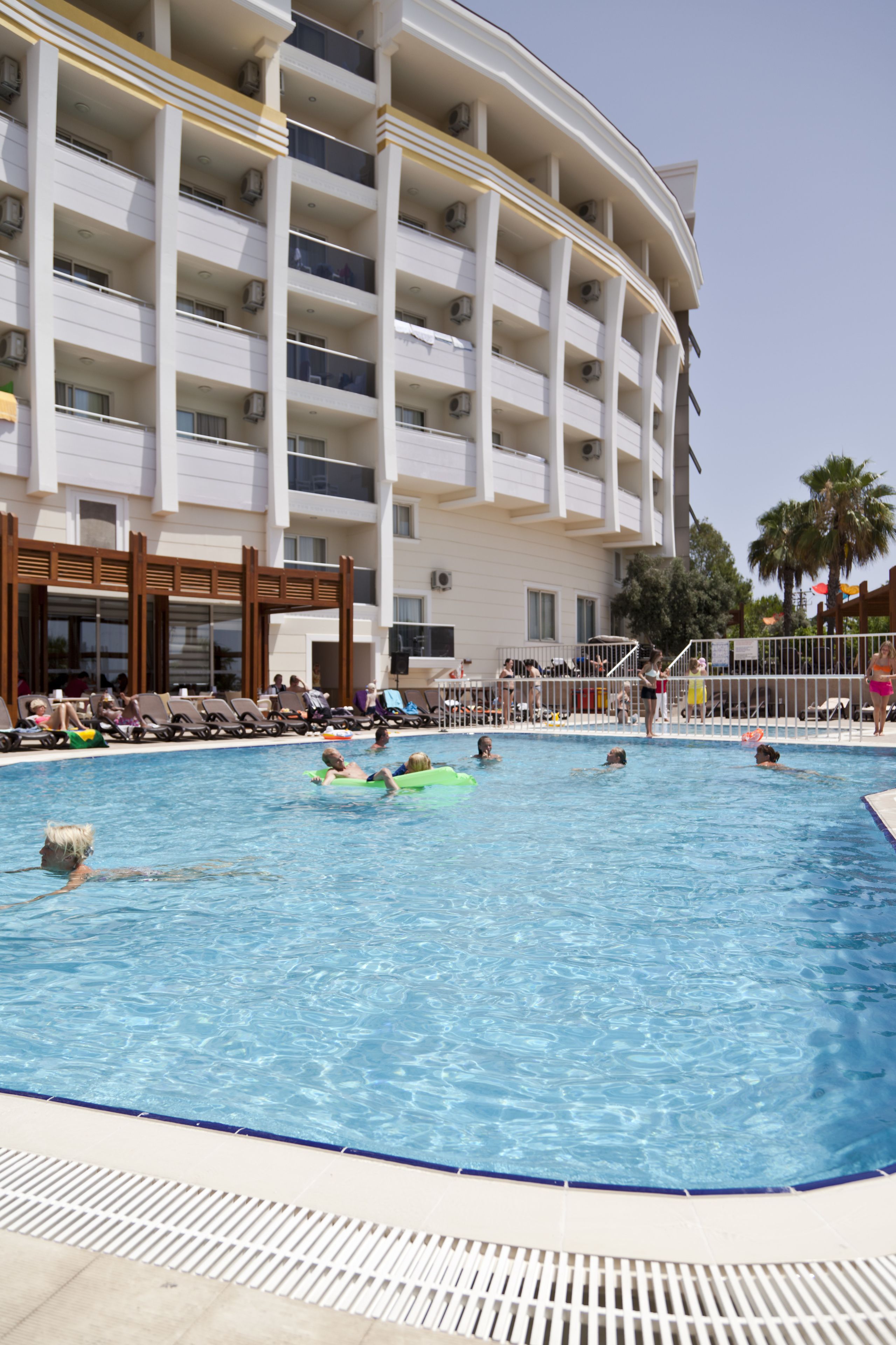 A swimming pool with people, a building with balconies, and a palm tree.