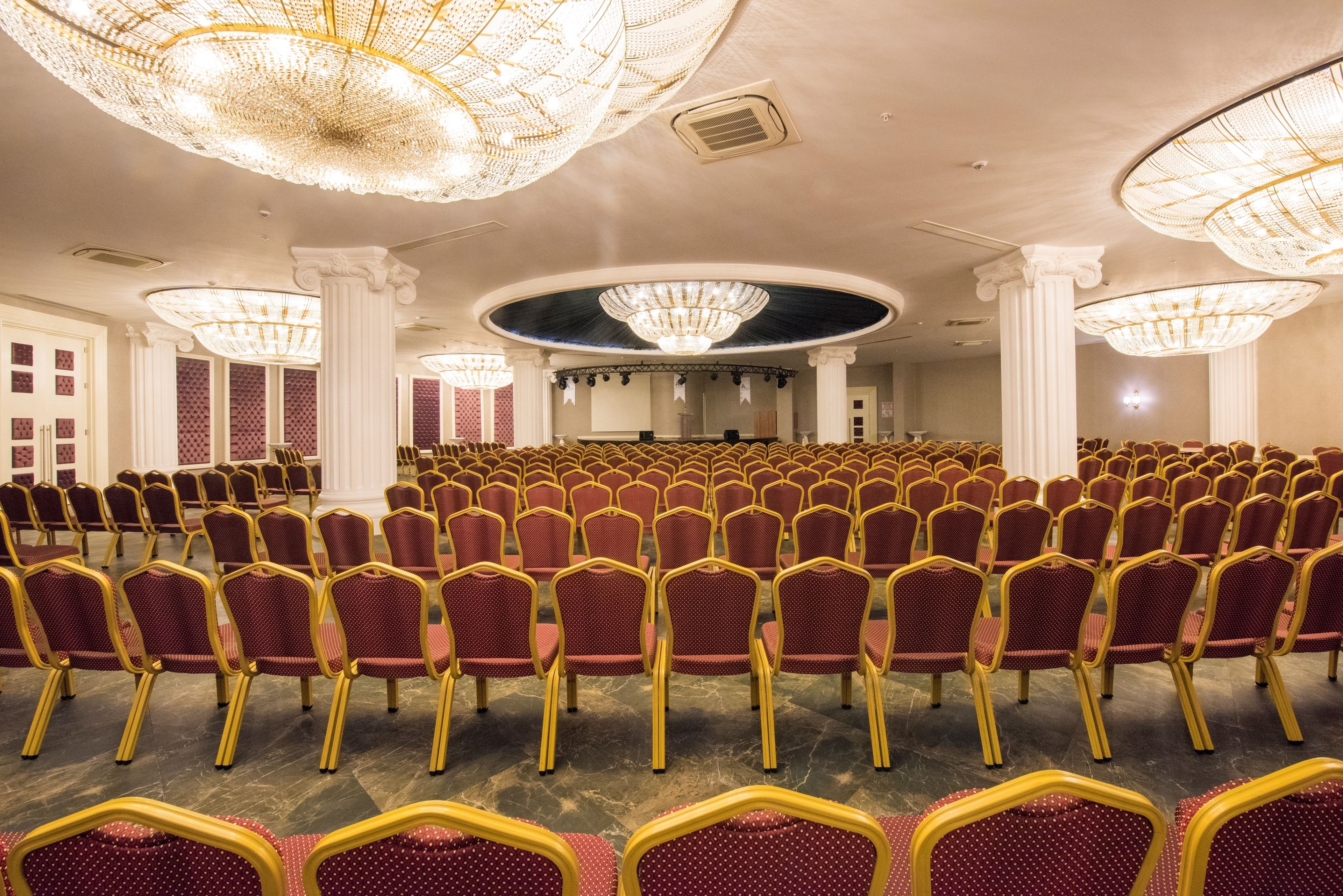 A large hall with rows of red chairs and gold frames, ornate chandeliers, and classical columns.