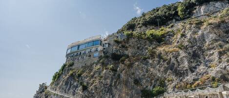 A coastal hotel with a stone facade, a swimming pool, and a beach with umbrellas and lounge chairs.
